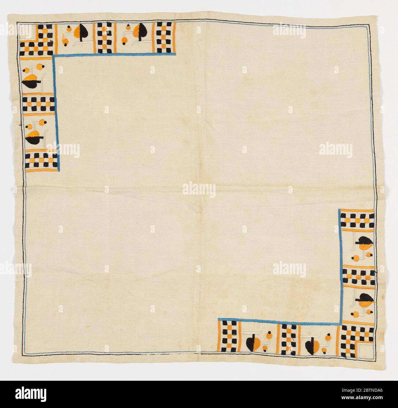 Table scarf. Square white tablecloth with L-shaped bands of embroidery in opposite corners. Design consists of spade-like leaves and berries that alternate with checkerboard bands in dark yellow and black embroidery with an inner blue border. Thin blue and black outer border. Stock Photo