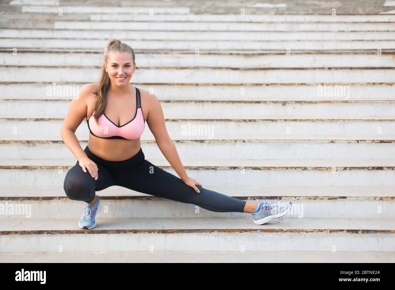 Young beautiful plus size woman in sporty top and leggings with red  headphones sitting on knees on yoga mat while thoughtfully looking aside in  city park Stock Photo
