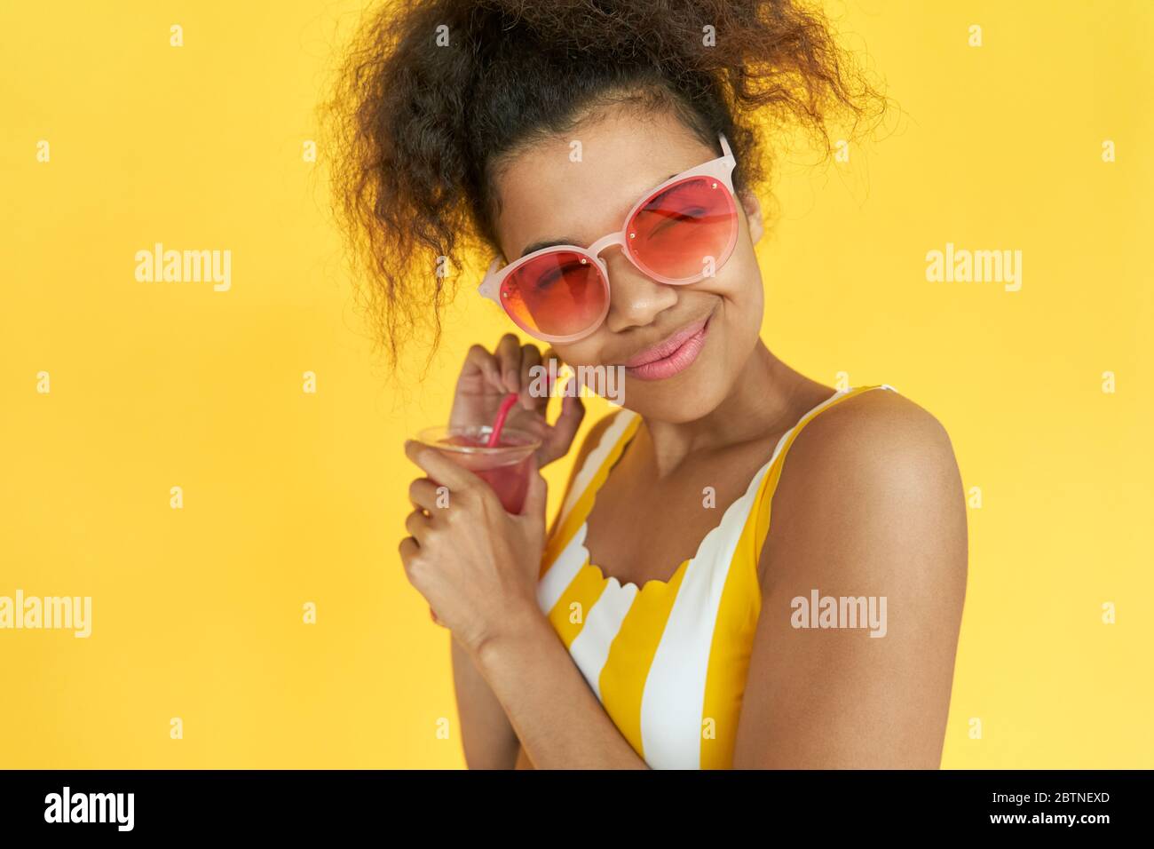 Happy african girl wear sunglasses hold drink wink at camera in yellow studio. Stock Photo