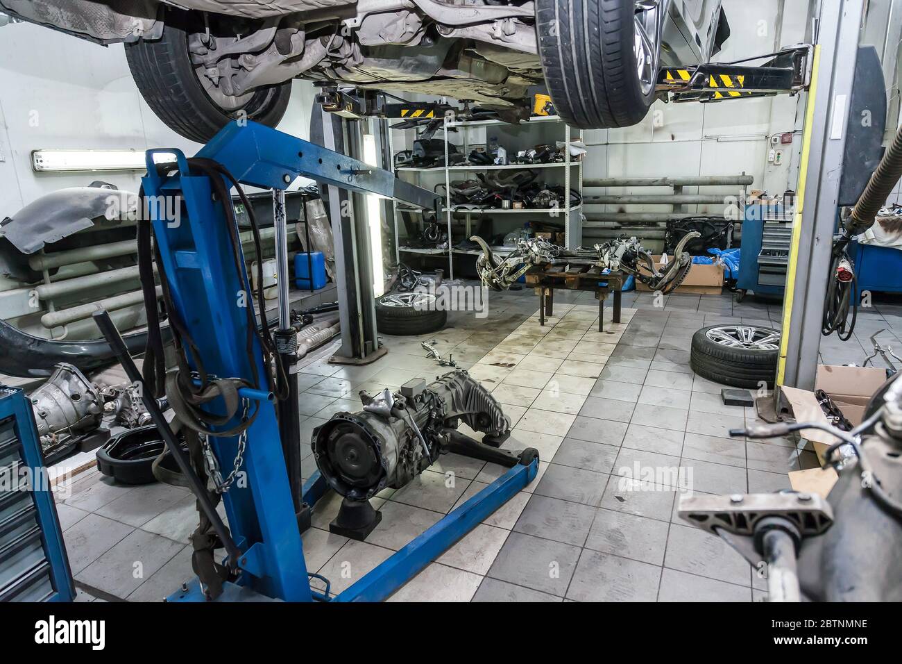 A raised car on a blue crane for engine or gearbox repair in a car workshop against the background of removed spare parts - wheels, rear axle axis. Stock Photo