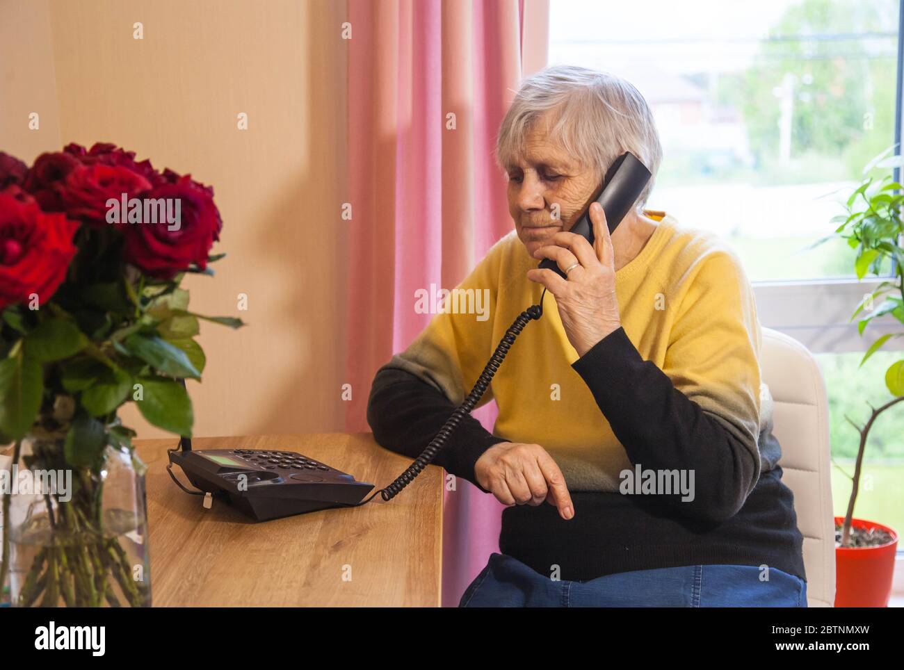 Grandmother is on the phone sitting in a chair in a cozy room with flowers Stock Photo