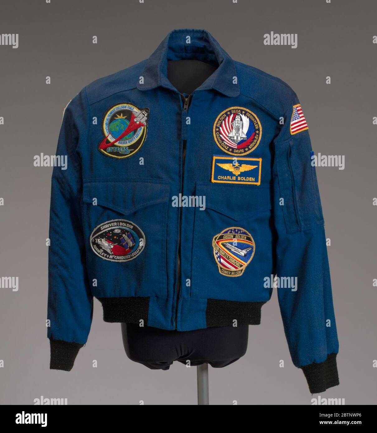 NASA flight jacket owned by Charles Bolden. Blue flight jacket featuring seven different patches. An American flag patch appears on the proper left shoulder of the flight jacket. A patch on the proper right shoulder depicts a space shuttle ascending vertically above stars set against a red, orange, and yellow background. Stock Photo