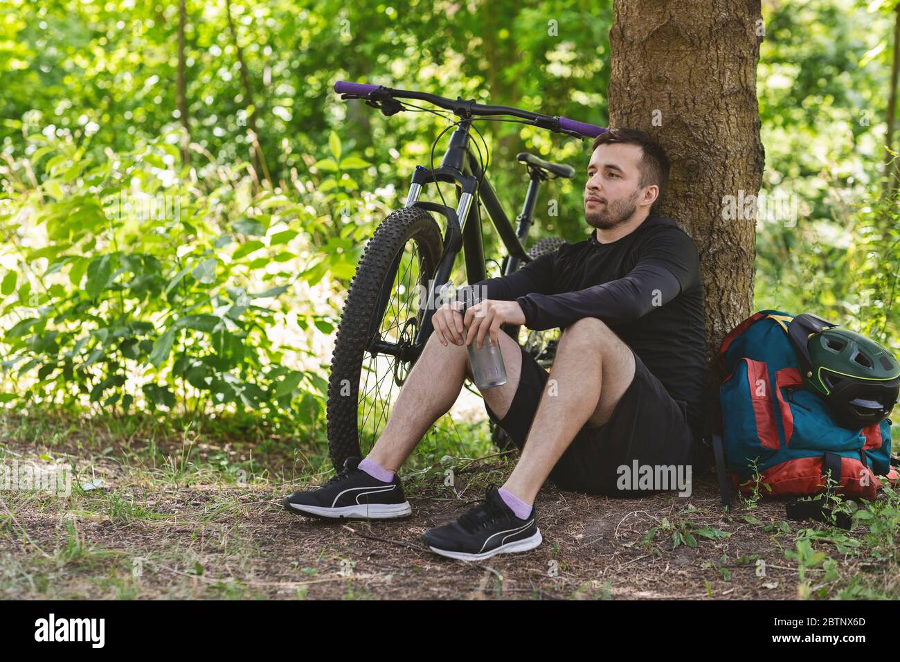 Joyful cyclist sitting under tree, drinking water Stock Photo