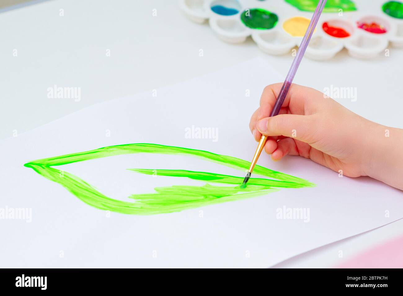 Child is drawing green leaf by watercolors on white paper. Earth day concept. Stock Photo