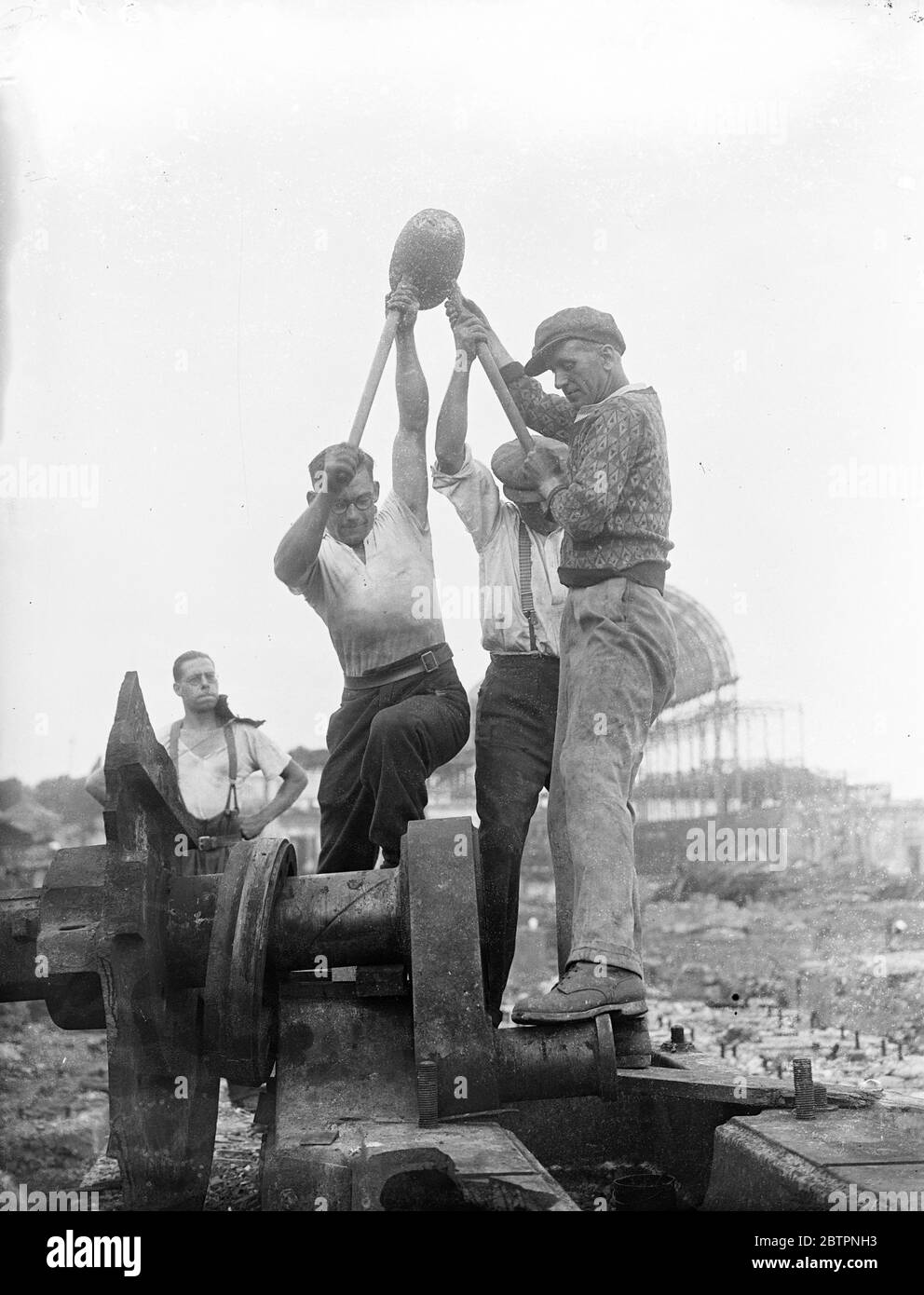 Three hands for this job. Waking up Crystal Palace with 100pound hammer. The huge hammer with a double shaft, weighing 100 pounds, and needing to men to wield it is being used in breaking up the debris left at Crystal Palace after the disastrous fire last year. Photo shows, three, workmen using the double shaft, 100 pound hammer at the Crystal Palace. 22 June 1937 Stock Photo
