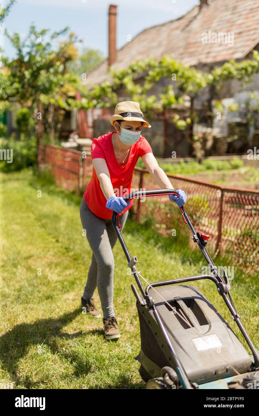 A woman in her backyard mowing grass with a lawn mower on a sunny day at  home wearing a surgical mask because of the coronavirus epidemic Stock  Photo - Alamy