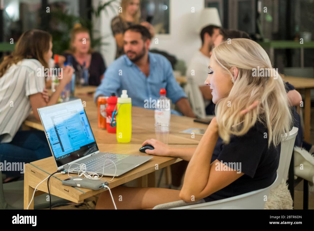 A woman works on a laptop while her co-workers meet and socialize in the background. Stock Photo