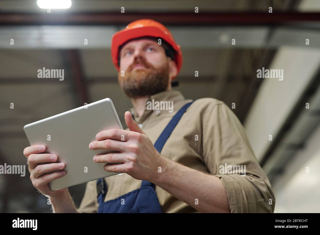 Below view of young bearded engineer in hardhat using tablet while controlling work at factory Stock Photo