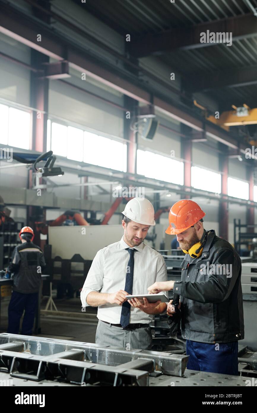 Technician in formalwear using tablet while engineering industrial machine with factory worker in workshop Stock Photo
