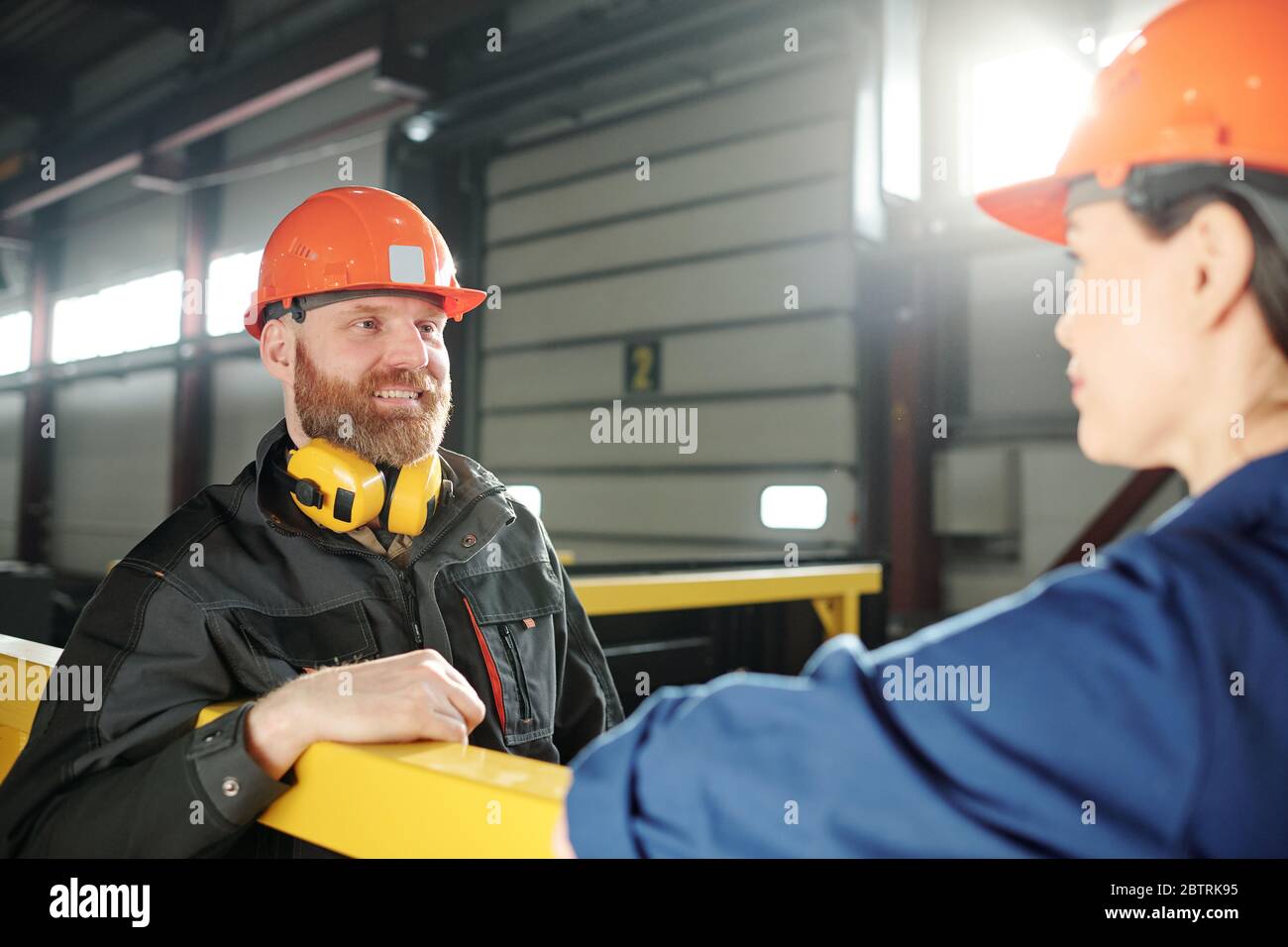 Cheerful handsome wormer in hardhat standing at railing and chatting with colleague during break at factory Stock Photo