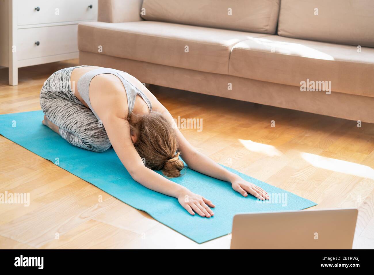 Young attractive women doing yoga exercise at home, Balasana, Child Pose, Ardha-Kurmasana Half Tortoise Pose in livingroom. Working out wearing sportswear bra and pants. Healthcare concept Stock Photo