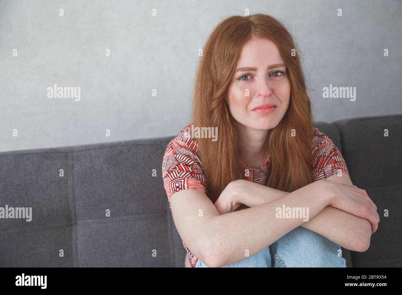 Young sad depressed caucasian woman portrait with tears in her eyes sitting on sofa clasping your knees Stock Photo