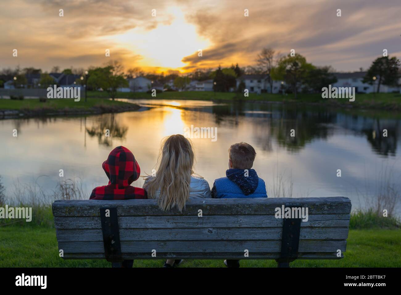 Family, mom with her two kids from back watching the sunset, sitting on a bench near a pond Stock Photo