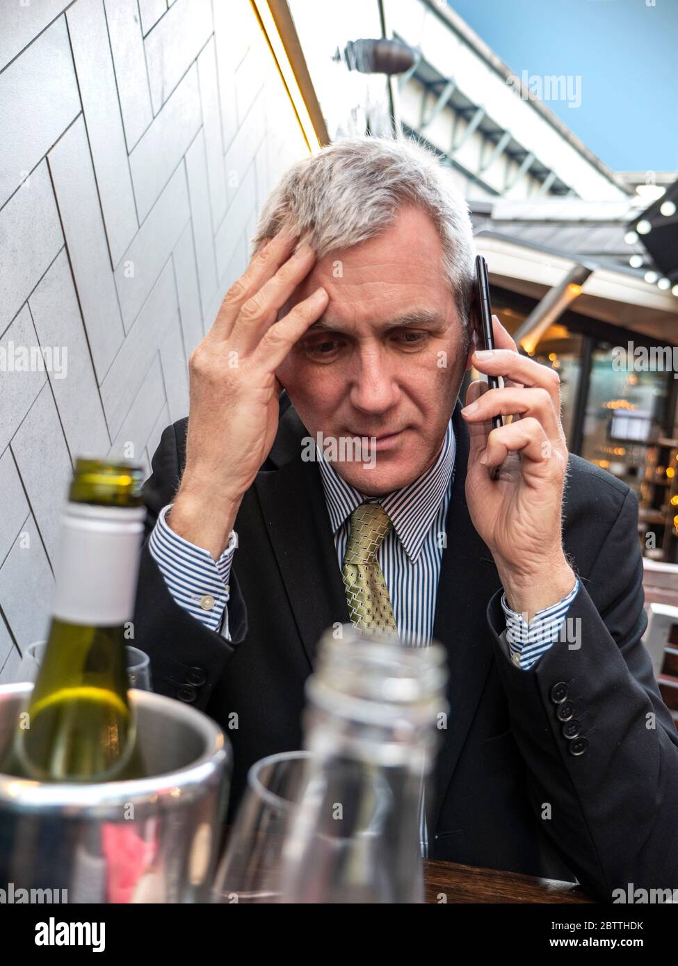 Businessman with alcoholic drinks on table looking worried, bad news, pensive, concerned, listening on his smartphone iPhone mobile telephone Stock Photo