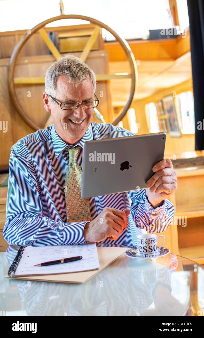 Zoom meeting in progress, mature businessman smiling in his barge boat office holding smart tablet Apple iPad computer in a virtual meeting situation Stock Photo