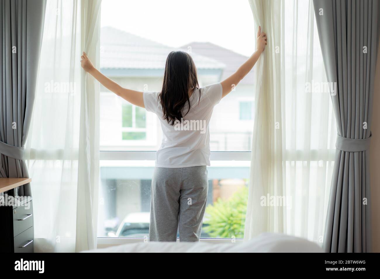 Rear view of Asian woman waking up in her bed fully rested opening window curtains and looking through the window in bedroom at home. Stock Photo