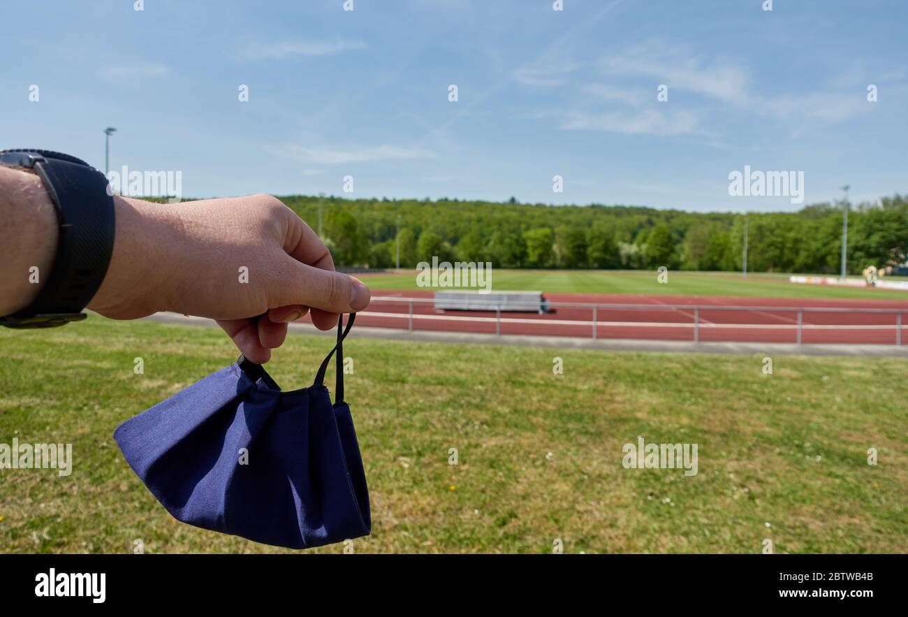 Mouth and nose mask for corona protection, is held by a large human hand. In the background, empty sports grounds in Germany. Stock Photo