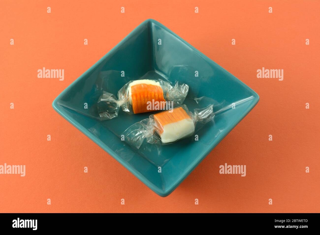 Two orange and champagne flavored chew candy pieces in plastic wrappers in blue square bowl on orange background Stock Photo