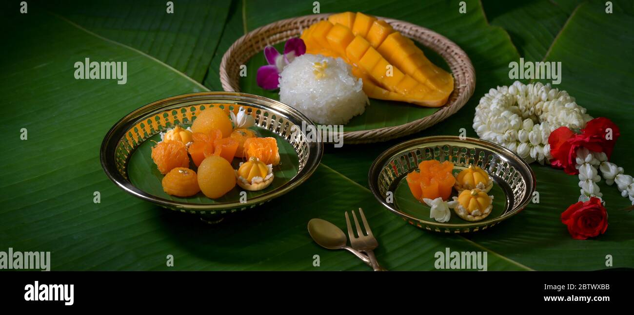 Cropped shot of several Thai traditional dessert serving on wooden tray and brass tray with brass cutlery on banana leaves background Stock Photo