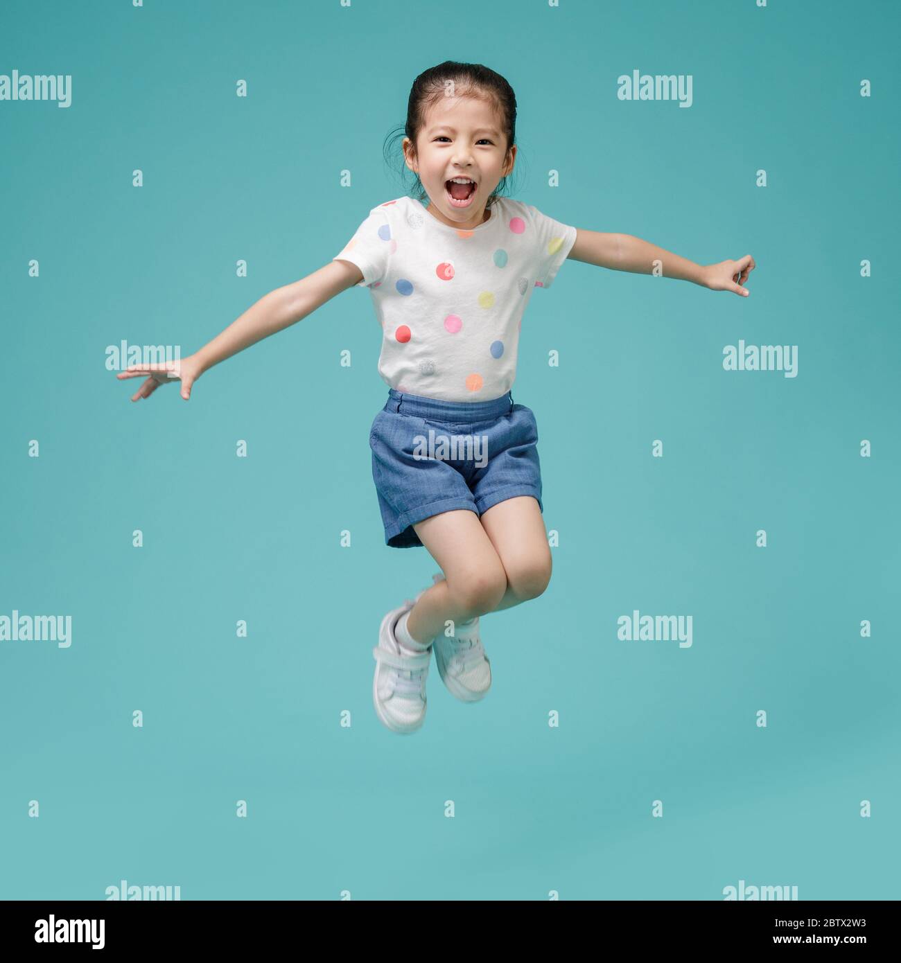 Playful energetic Asian little girl jumping in mid-air, empty space in studio shot isolated on colorful blue background Stock Photo