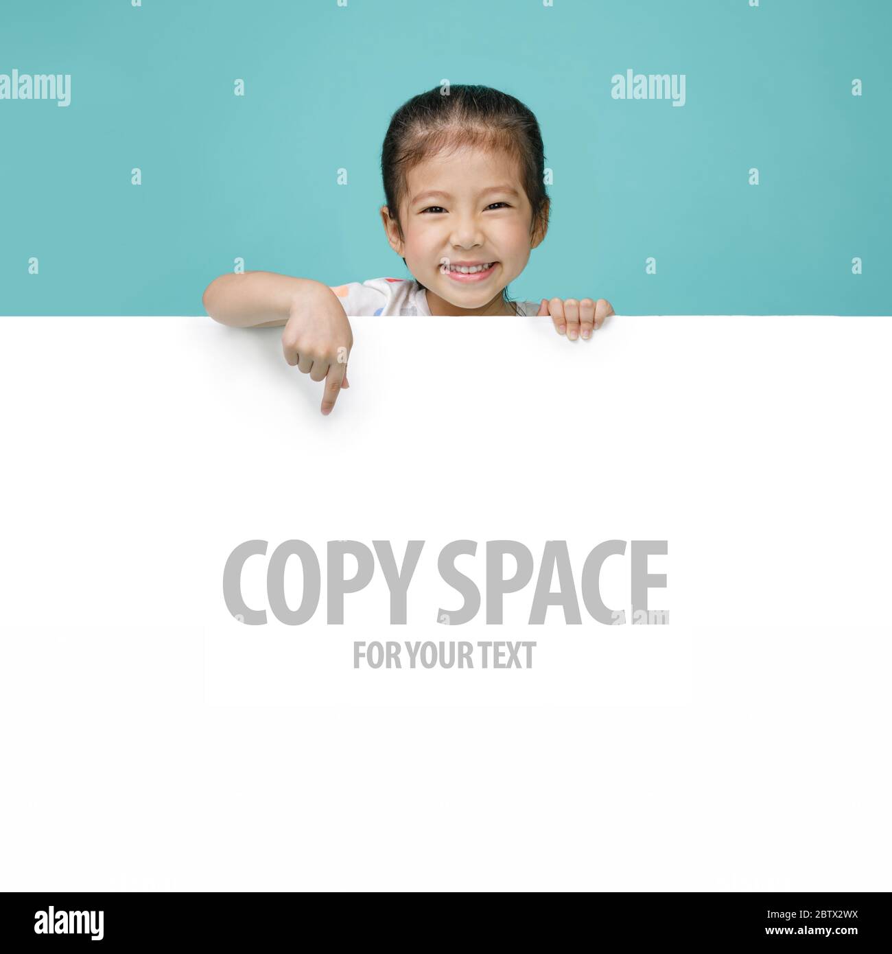 Excited Asian little girl pointing hand behind a blank white board, empty space in studio shot isolated on colorful blue background Stock Photo