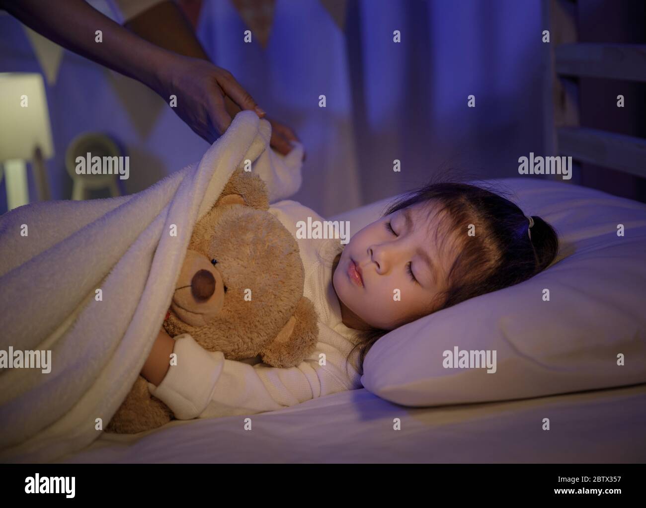 Mother putting blanket on and stroking head for her little girl daughter on bed in a dark bedroom at night, Child asian girl hug teddy bear, Comfortab Stock Photo