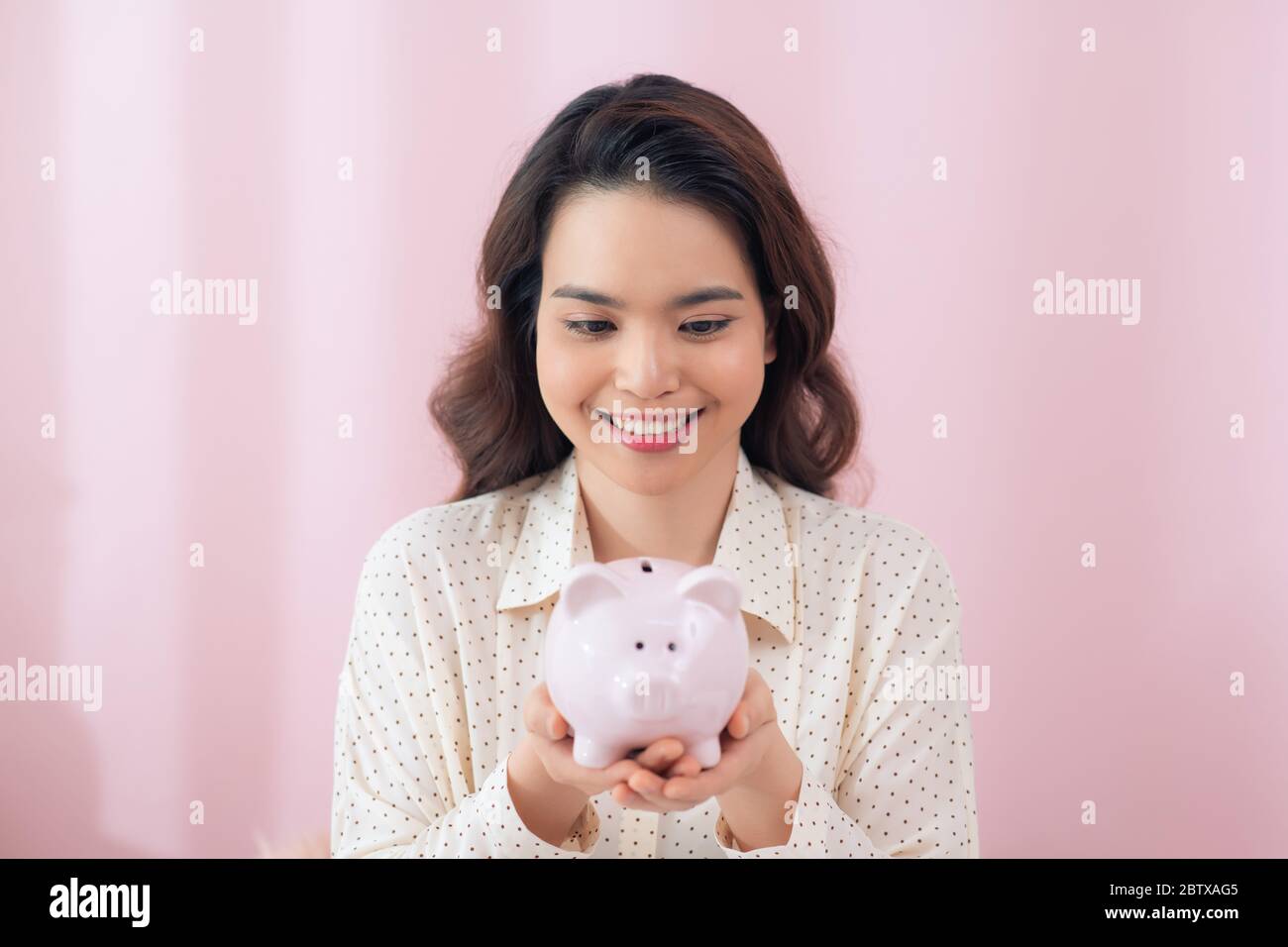 Beautiful young Asian woman holding piggy bank over pink background ...