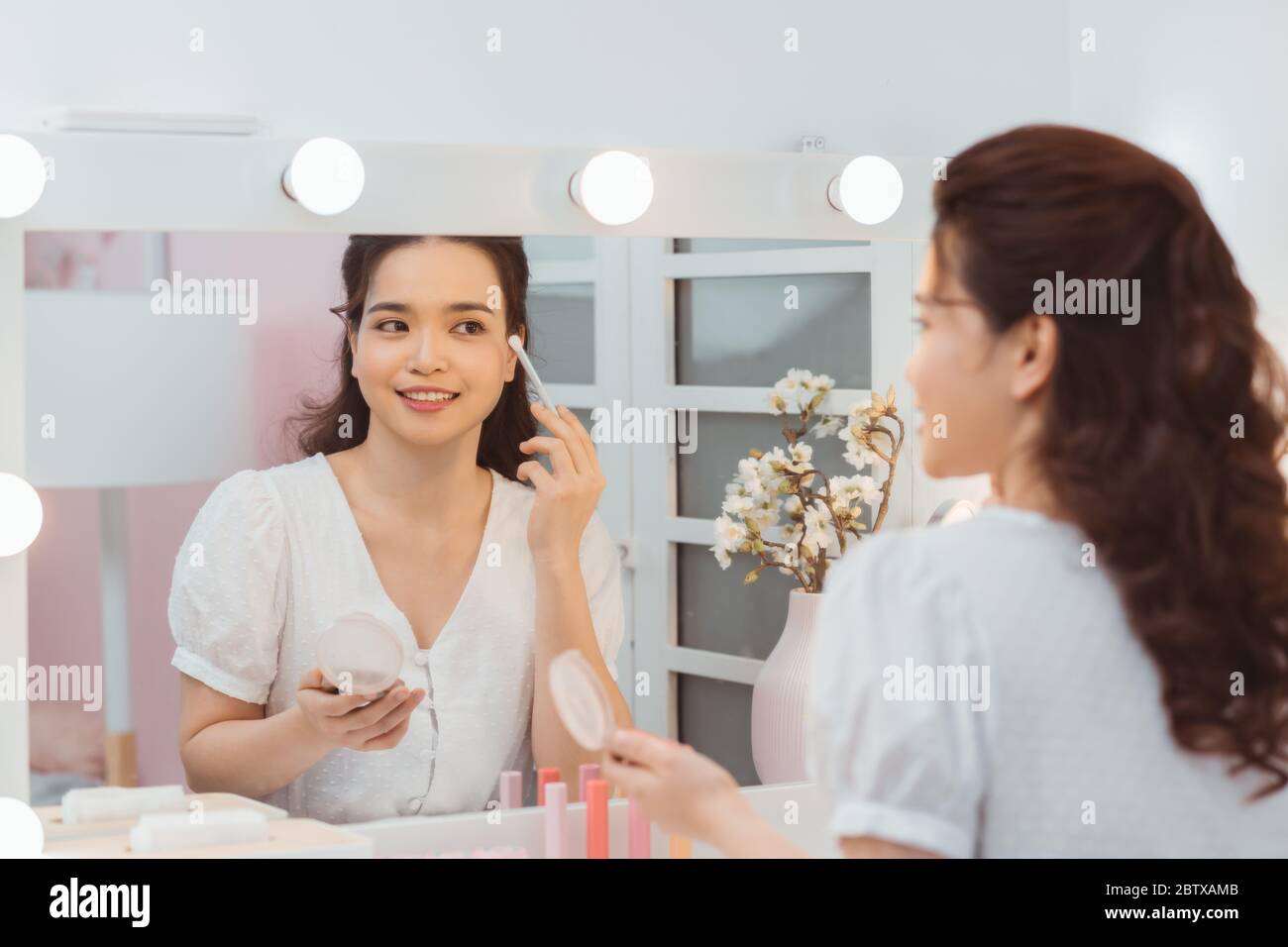 Portrait view of young adult asian woman looking at mirror, sitting in bright room and making make up. Beautiful and professional make up artist using Stock Photo