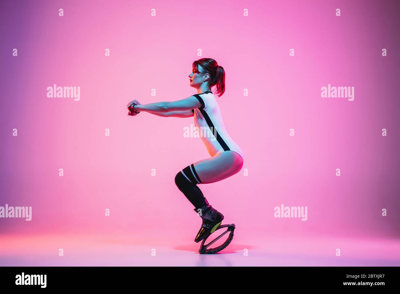 Beautiful redhead woman in sportswear jumping in a kangoo jumps shoes  isolated on purple-pink gradient studio background in neon light. Active  movement, action, fitness and wellness. Fit female model Stock Photo -