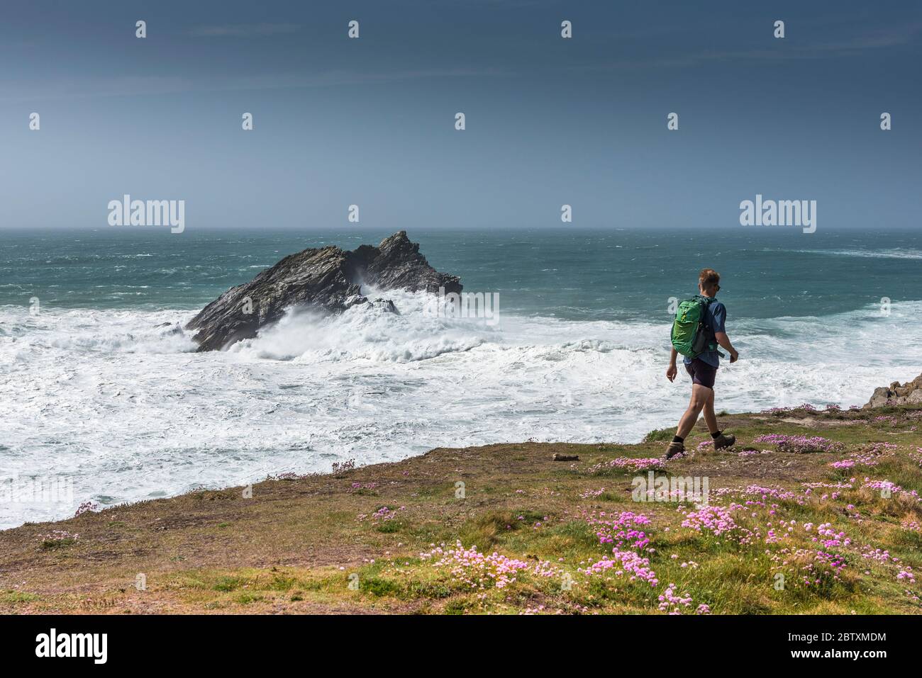 A walker on the coast path as wild waves break over The Goose an uninhabited rocky island off the coast of Pentire Point East in Newquay in Cornwall. Stock Photo