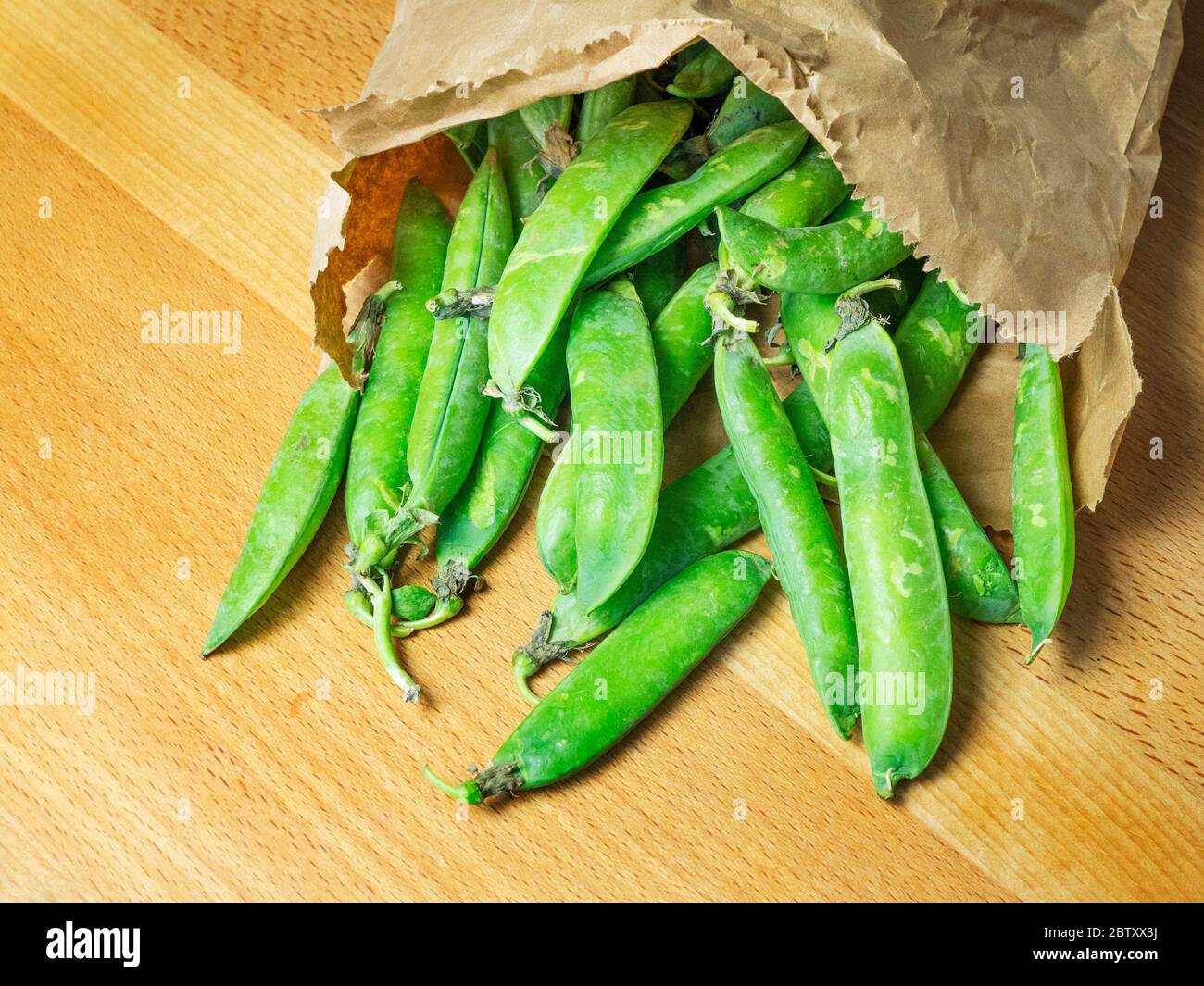 A paper bag of fresh garden peas in pods spilling onto a wooden kitchen table Stock Photo