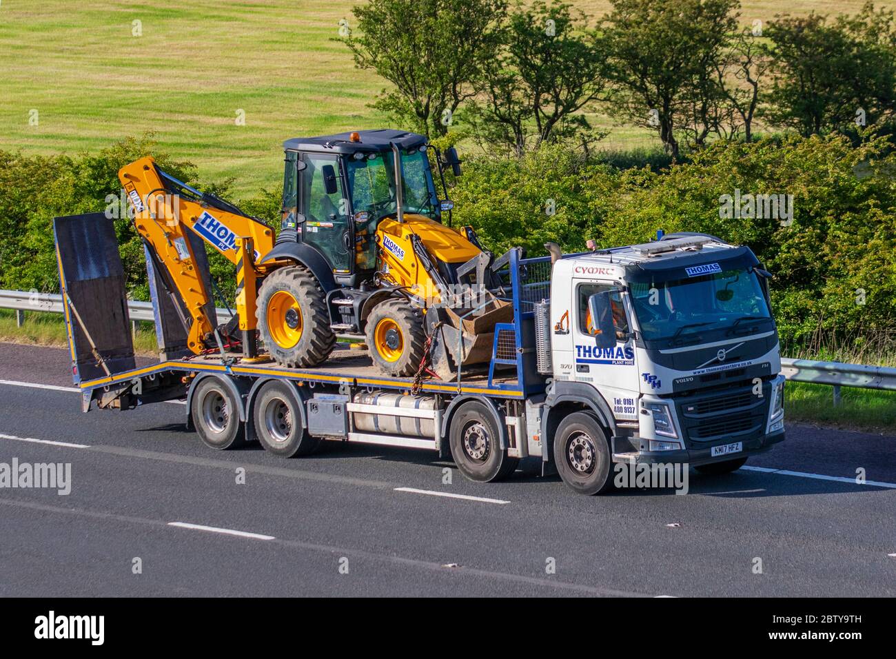 Thomas Plant Hire Wales; Haulage delivery trucks, lorry, transportation, truck, trailer carrying JCB 3CX backhoe loader, lowloader cargo carrier, Volvo vehicle, European commercial transport industry HGV, M6 at Manchester, UK Stock Photo