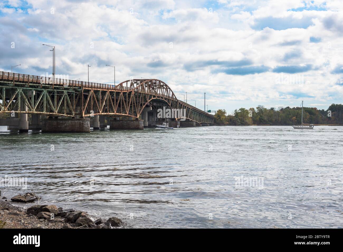 Boats sailing up a mighty river passing an old rusty steel road bridge on a cloudy autumn day Stock Photo