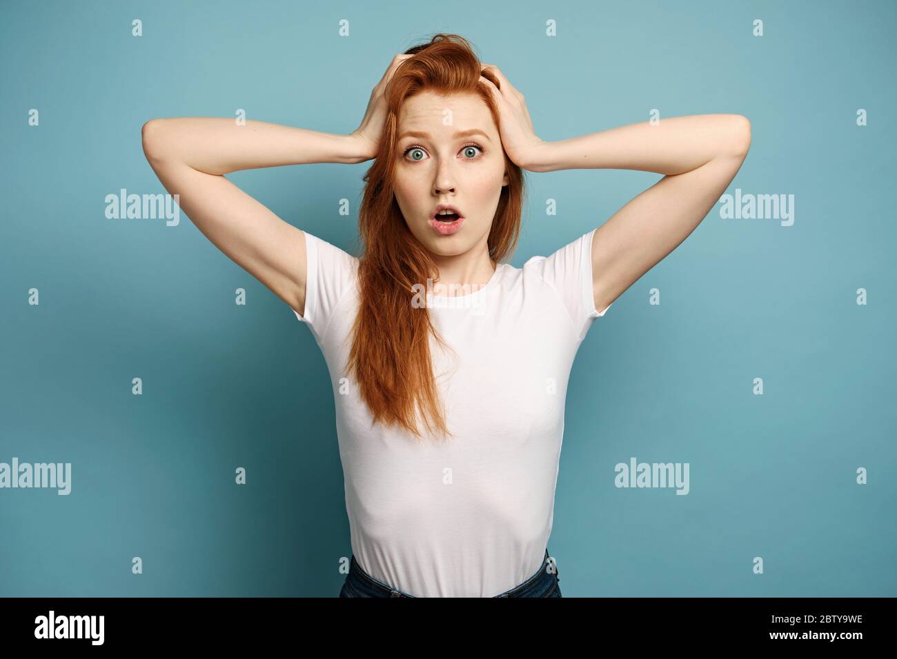 The red-haired girl stands on a blue background in a white T-shirt, in horror holds hands behind head, opening her mouth Stock Photo