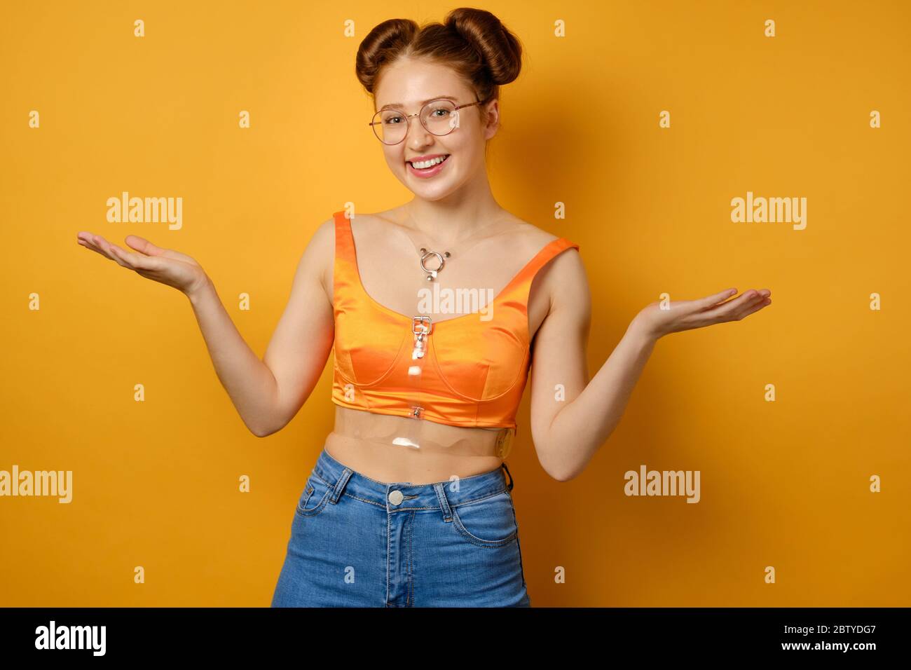 The girl with two beams, glasses and an orange top stands on a yellow background and smiles, spreading her palms to the sides Stock Photo