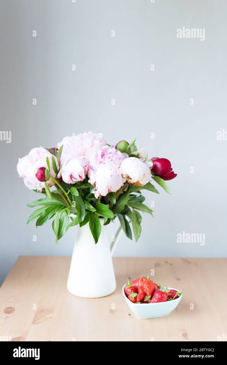 Bunch of Pink peonies in vase and strawberry on the wooden table . Flowers on a beige wooden table near the window. Home house interior. Beautiful Stock Photo