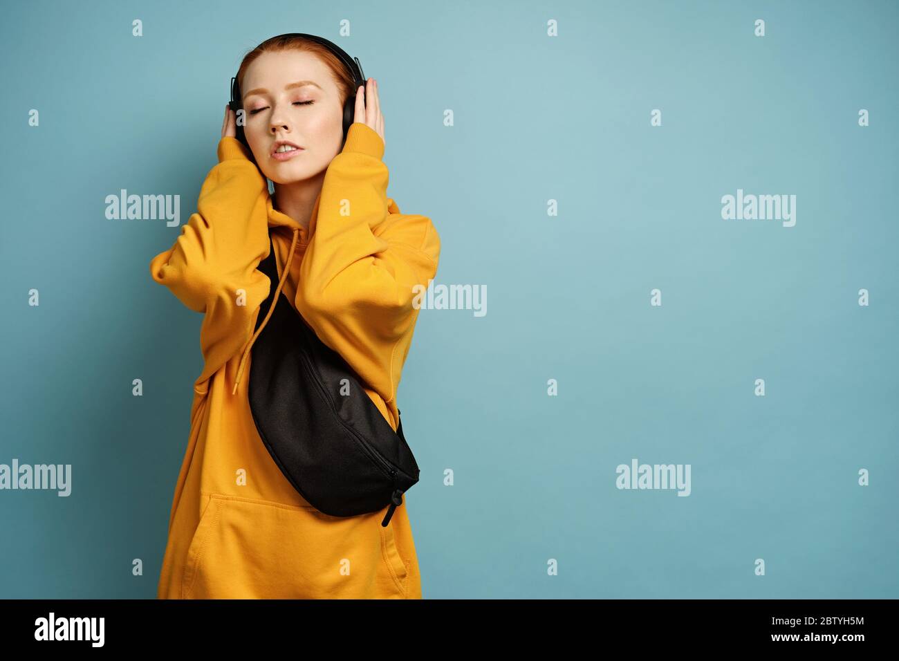 Red-haired girl in a yellow sweatshirt stands on a blue background, closing her eyes and holding her hands on headphones Stock Photo