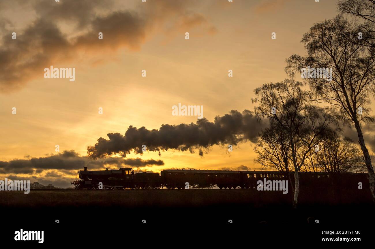 Side view of puffing vintage UK steam train silhouette, passing through Worcestershire countryside landscape in beautiful, evening winter sunset. Stock Photo
