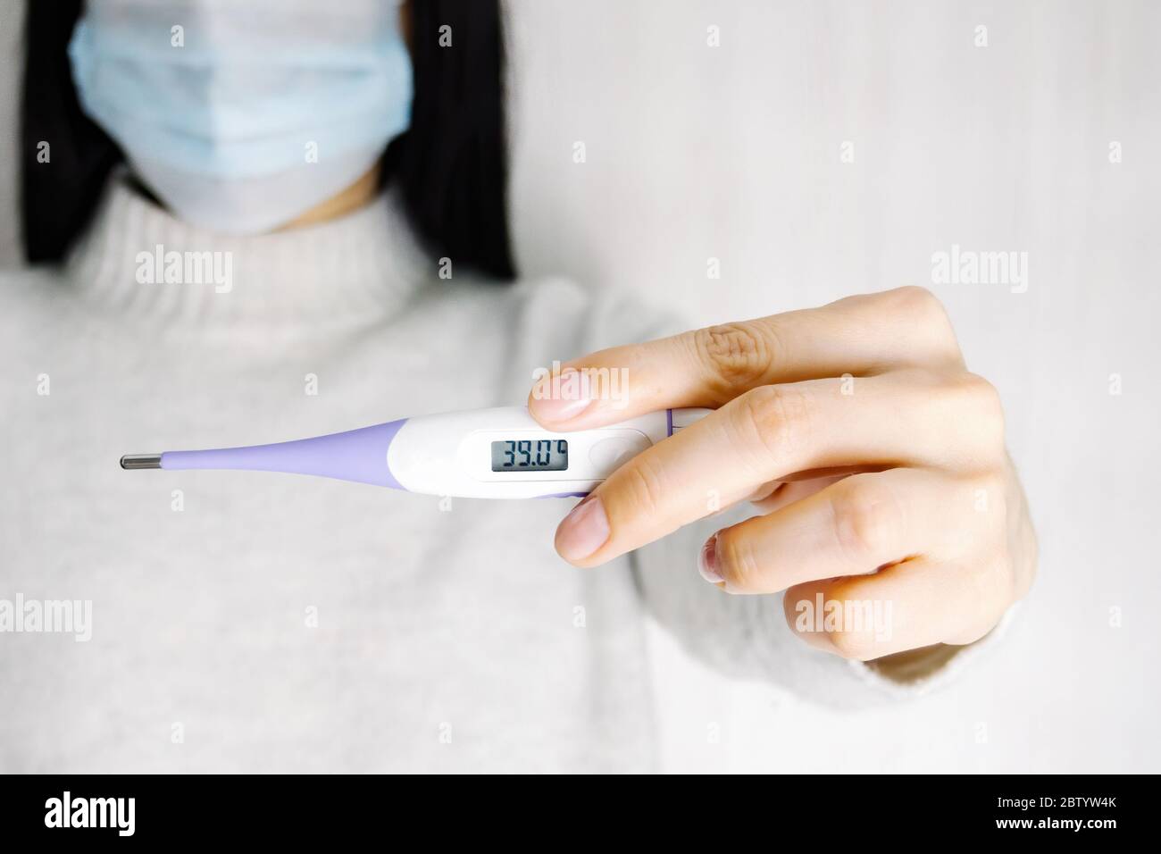 Young woman in medical disposable mask holds an electronic thermometer in her hands. High temperature on display, selective focus. Stock Photo