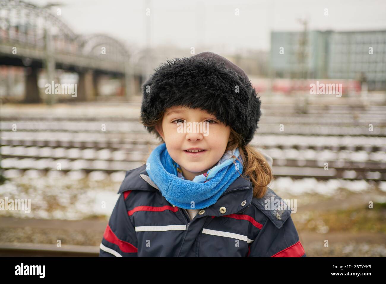 Adorable little girl in outerwear and hat smiling and looking at camera while standing on blurred background of rails and bridge on railroad station Stock Photo