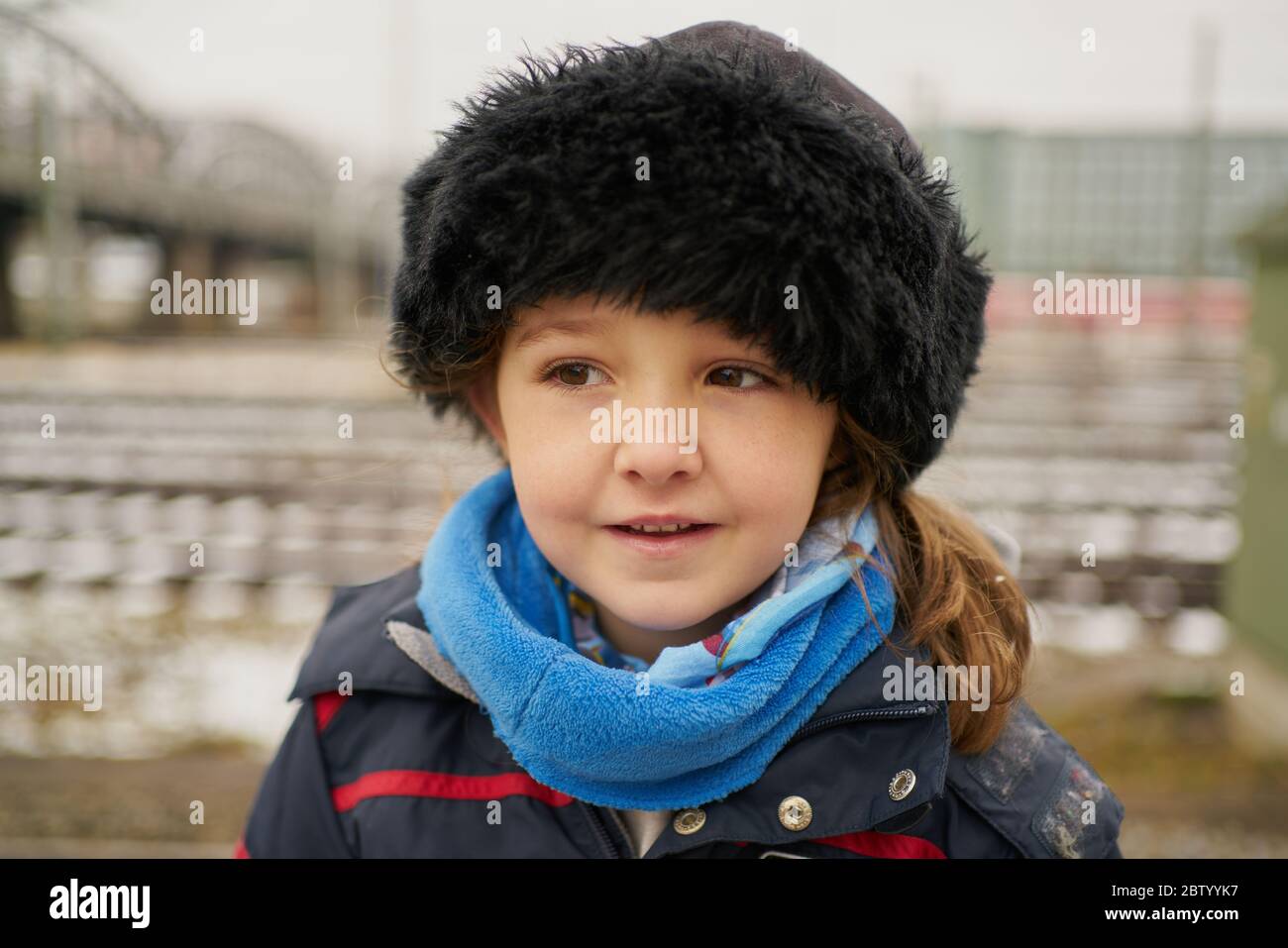 Adorable little girl in outerwear and hat smiling and looking at camera while standing on blurred background of rails and bridge on railroad station Stock Photo
