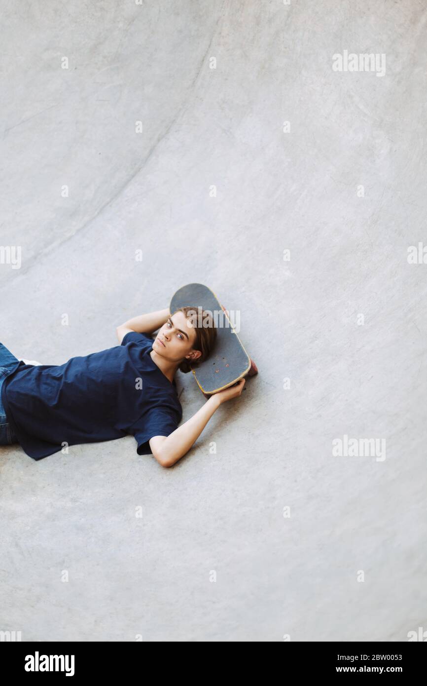 Cool young guy thoughtfully looking in camera while lying on skateboard in empty bowl at modern skatepark Stock Photo