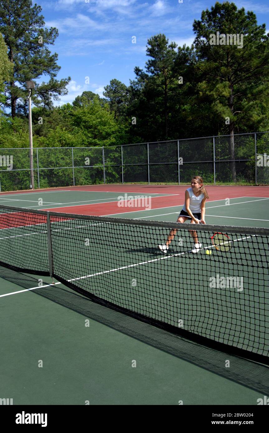 High school varsity tennis player hustles to the net but misses the tennis ball.  She is wearing a black tennis skrt and white tee shirt.  The court i Stock Photo