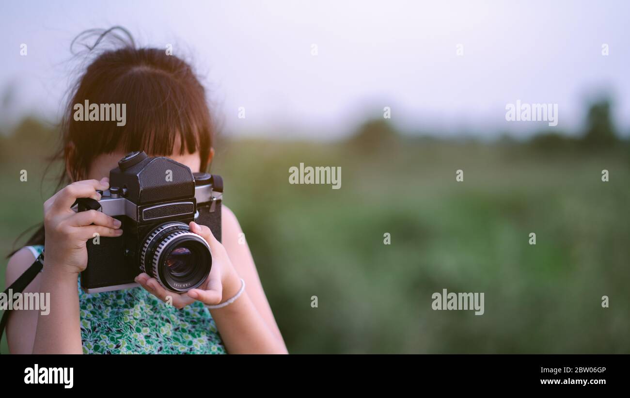 Little child girl holding medium format film camera and taking photo of sunset landscape with green field background Stock Photo