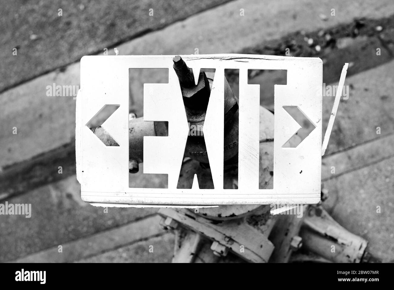 White Exit sign stuck on fire hydrant on sidewalk.  Abstract image of broken Exit sign in black & white horizontal photograph. Stock Photo