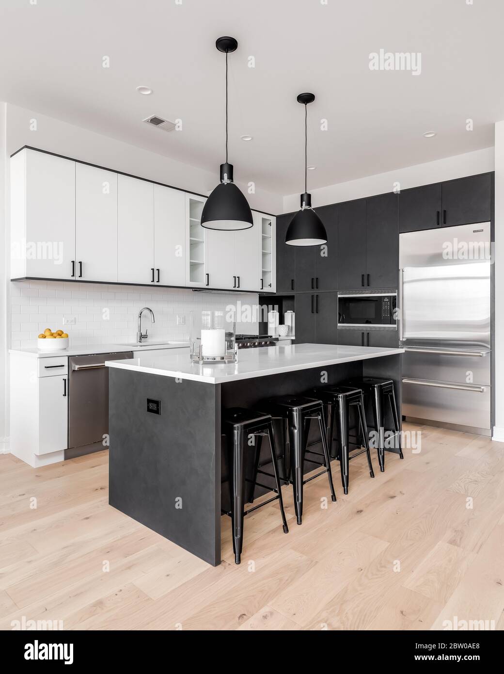 A modern kitchen with black and white cabinets, stainless steel Wolf and Sub-Zero appliances, and bar stools sitting at the white granite counter top. Stock Photo