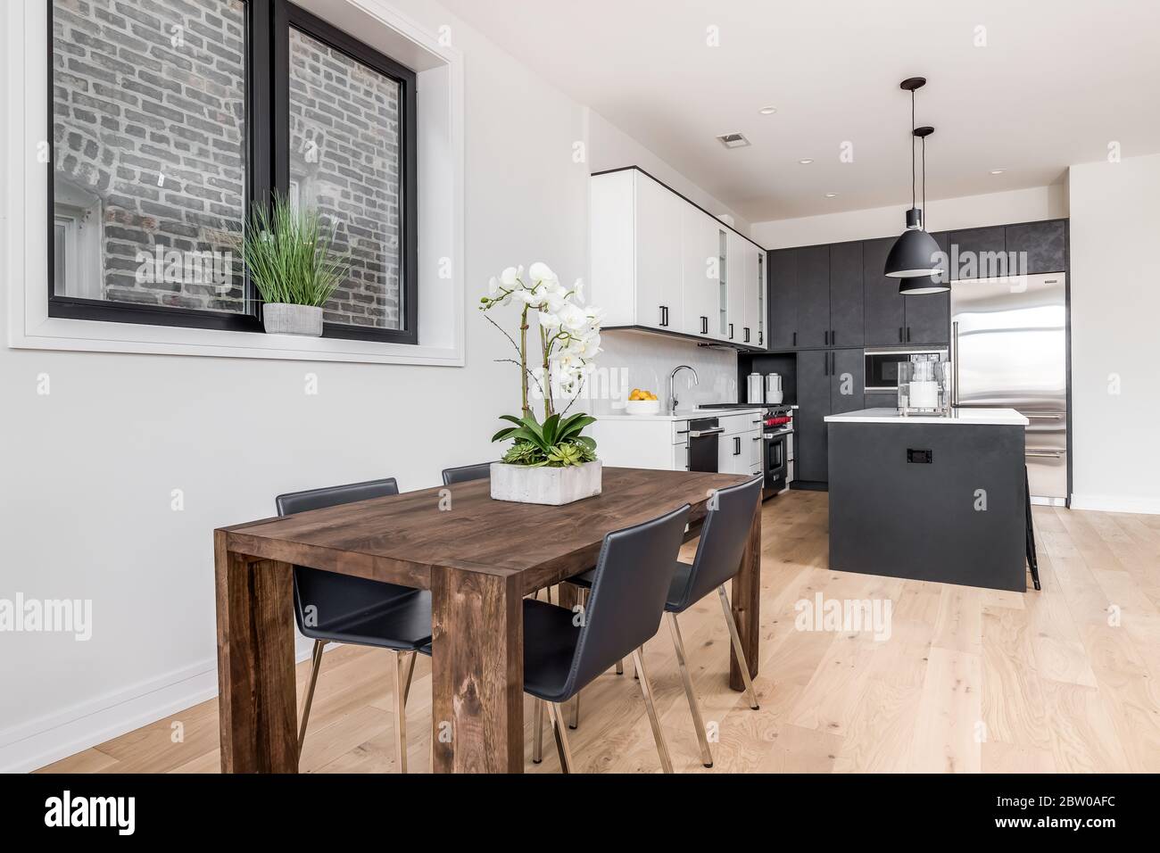 A dining room and kitchen with white and dark grey kitchen cabinets. A flower is on the table as a centerpiece with four chairs at the wooden table. Stock Photo