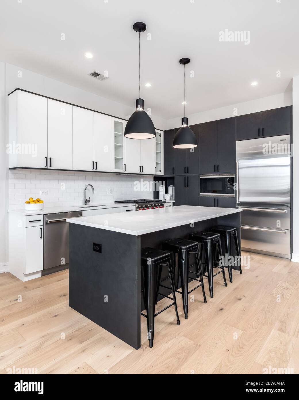 A modern kitchen with black and white cabinets, stainless steel Wolf and Sub-Zero appliances, and bar stools sitting at the white granite counter top. Stock Photo