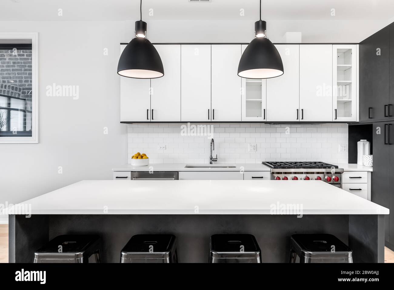 A modern kitchen with black and white cabinets, stainless steel Wolf and Sub-Zero appliances, and bar stools sitting at the white granite counter top. Stock Photo