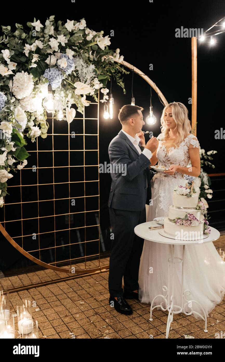 Beautiful newlywed standing near the wedding arch and tasting their three-tiered wedding cake decorated with flowers Stock Photo